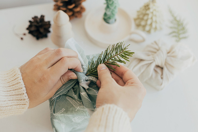 零浪费的圣诞理念。双手装饰礼盒包装在织物上的杉木枝白色木桌与环保装饰和蜡烛。Furoshiki礼品包装。Eco寒假图片下载