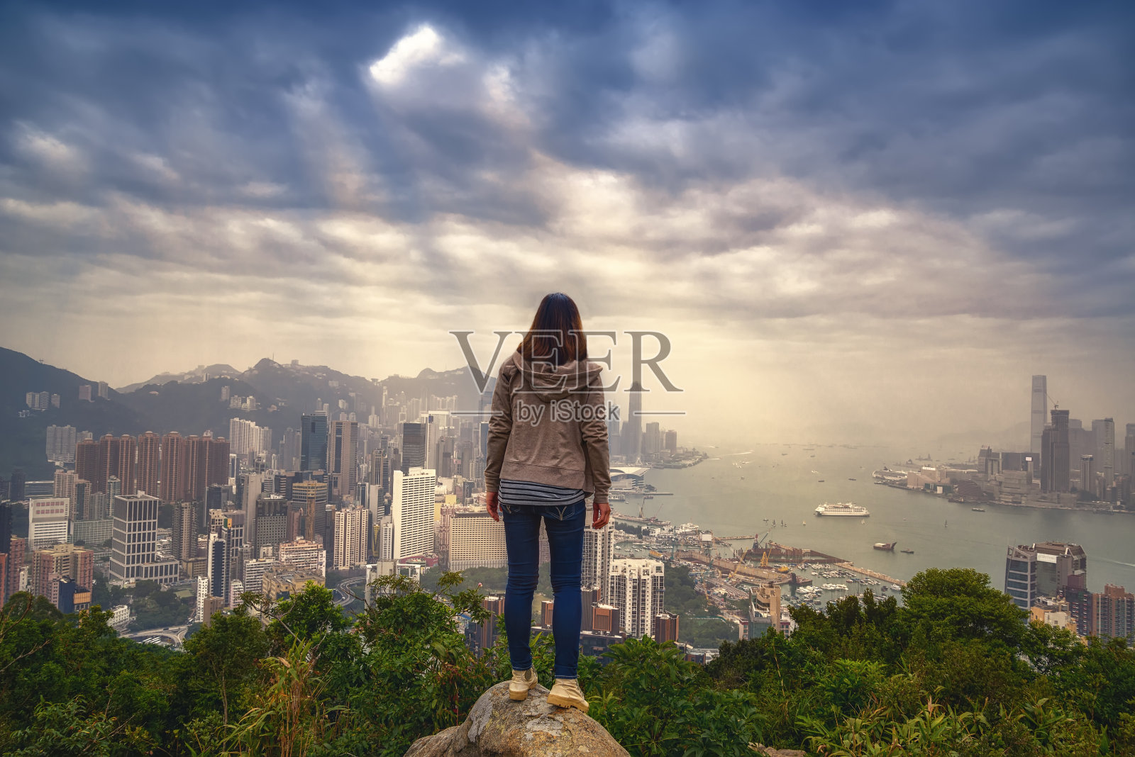 女孩与背包客享受阳光在山顶雾蒙蒙的城市。山顶上的岩石和日落在香港城市。成功,冠军,领袖的概念。照片摄影图片