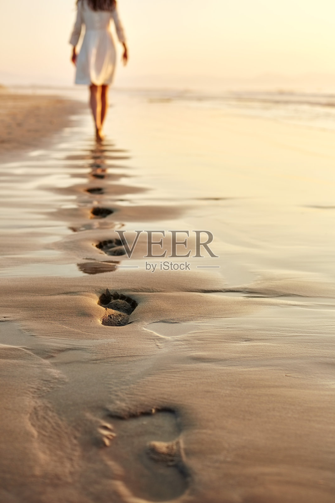 一个女人赤脚走在潮湿的岸边，在沙滩上留下脚印照片摄影图片
