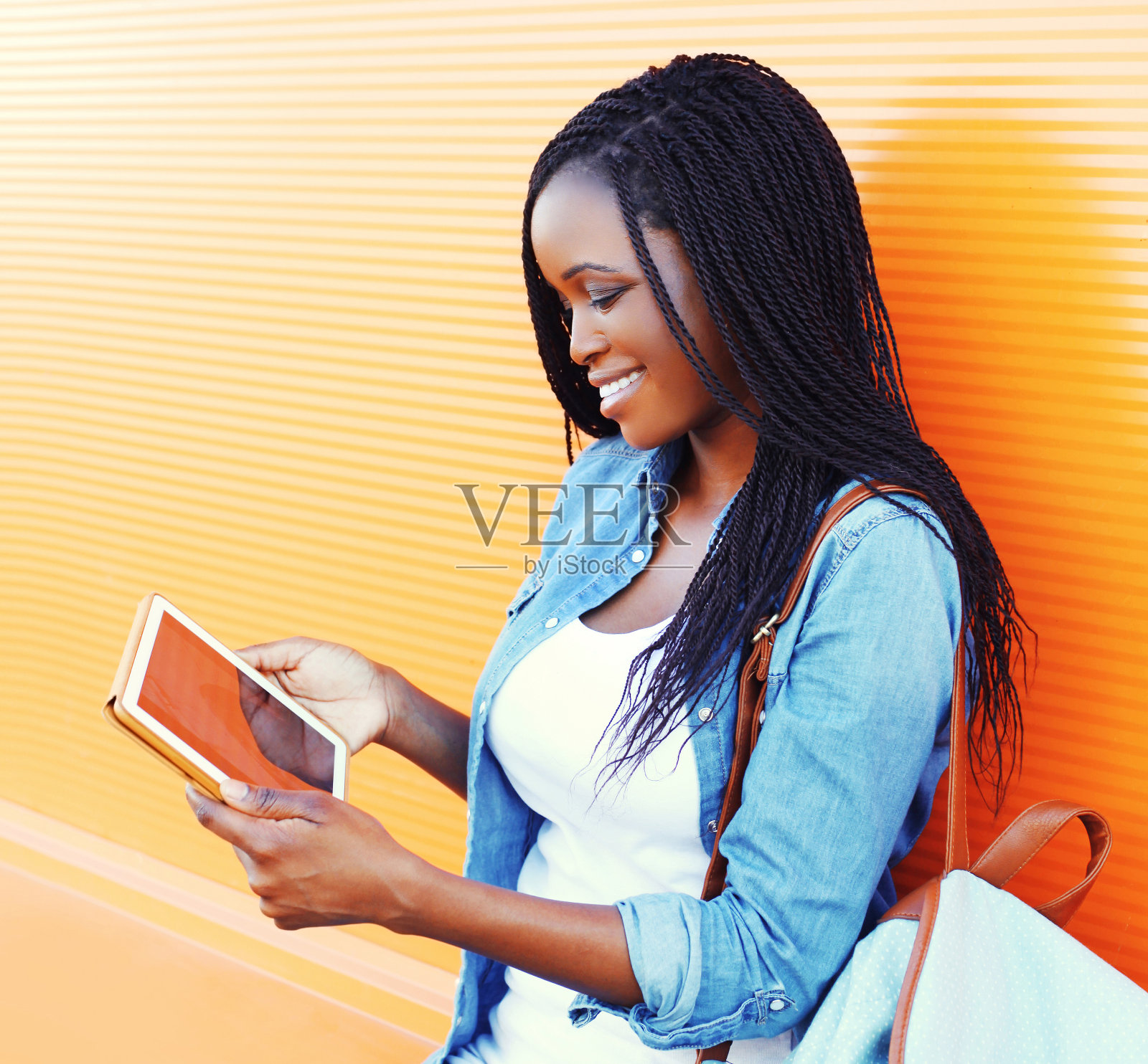 时尚年轻的非洲妇女微笑着使用平板电脑在城市在彩色的橙色背景照片摄影图片