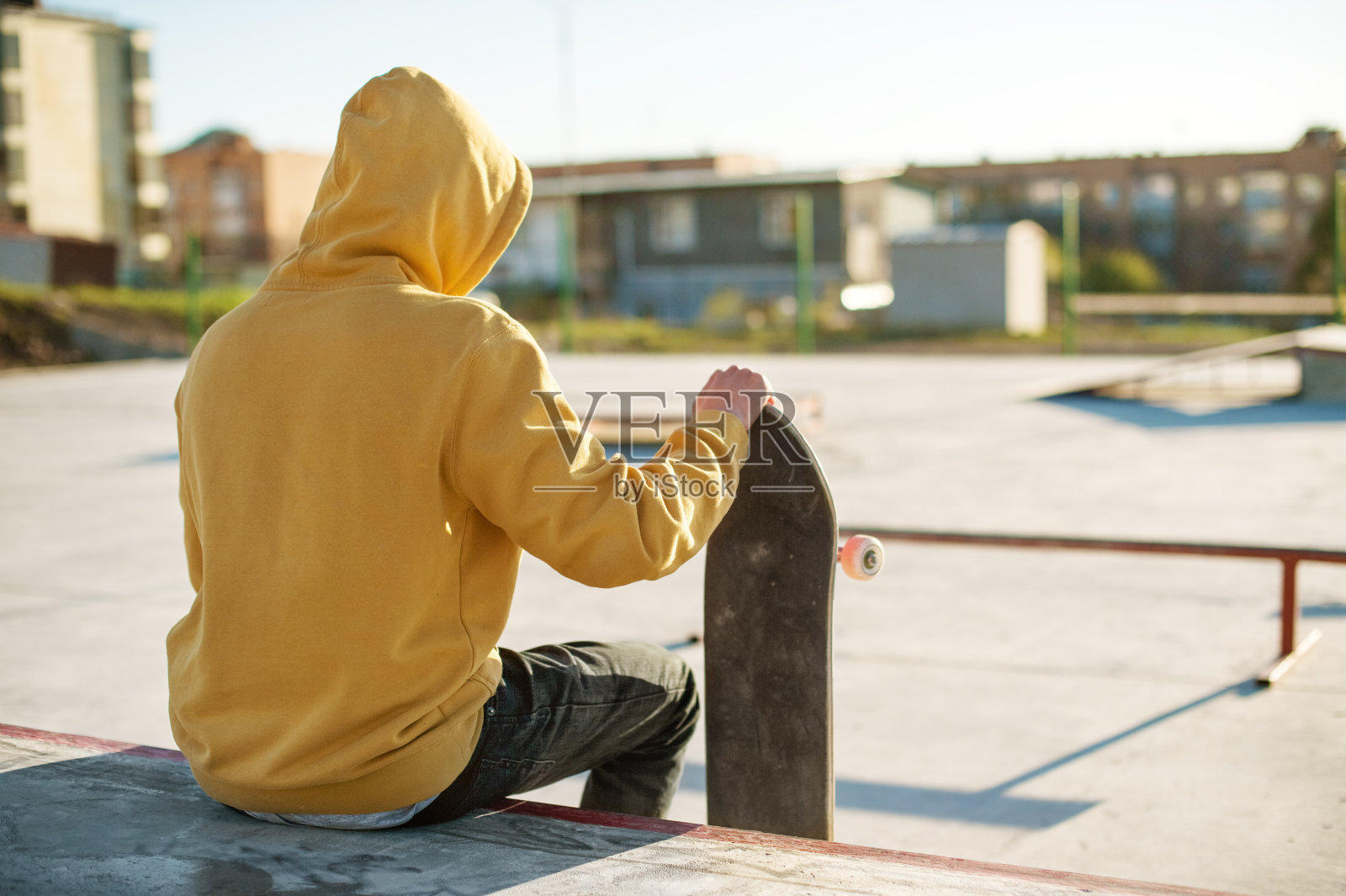 一个十几岁的穿着牛仔帽衫坐在一个滑板公园，拿着滑板的特写照片摄影图片