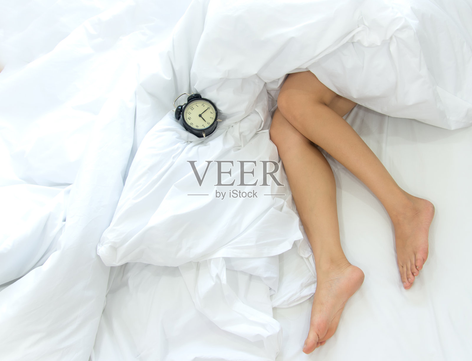 女性的腿和闹钟在床上的特写。懒惰的女人懒洋洋地躺在卧室里放松照片摄影图片