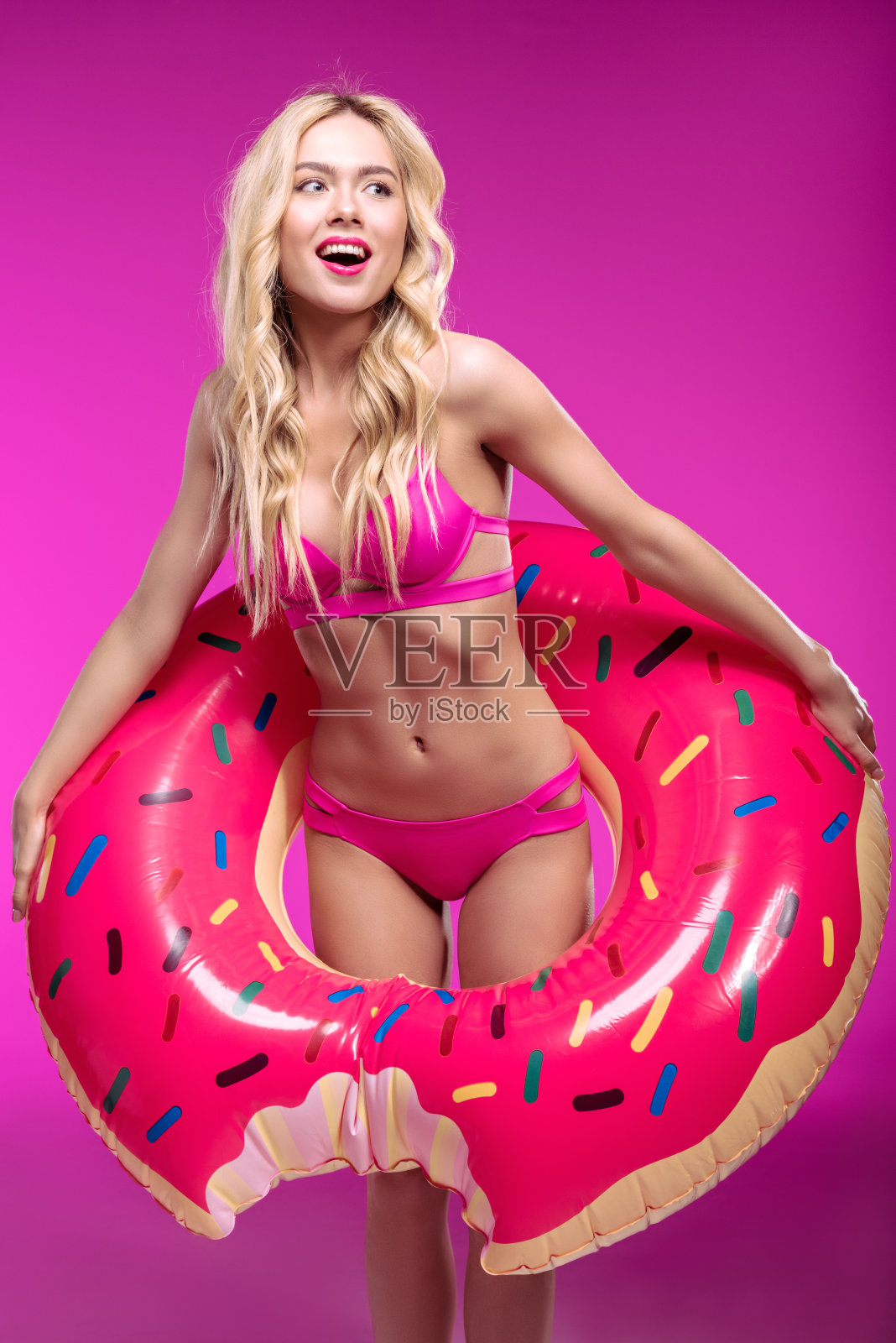 快乐的金发女人在比基尼穿着游泳管的形状的甜甜圈和看向别处照片摄影图片