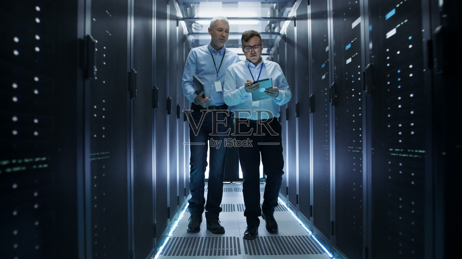 在数据中心，两个IT工程师穿过一排排的服务器机架。他们在平板电脑和笔记本电脑上工作。照片摄影图片