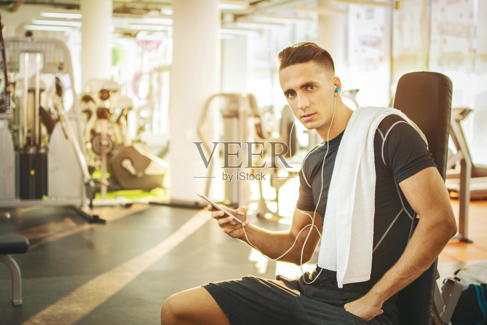 英俊的年轻人戴着耳机在健身房用智能手机。照片摄影图片