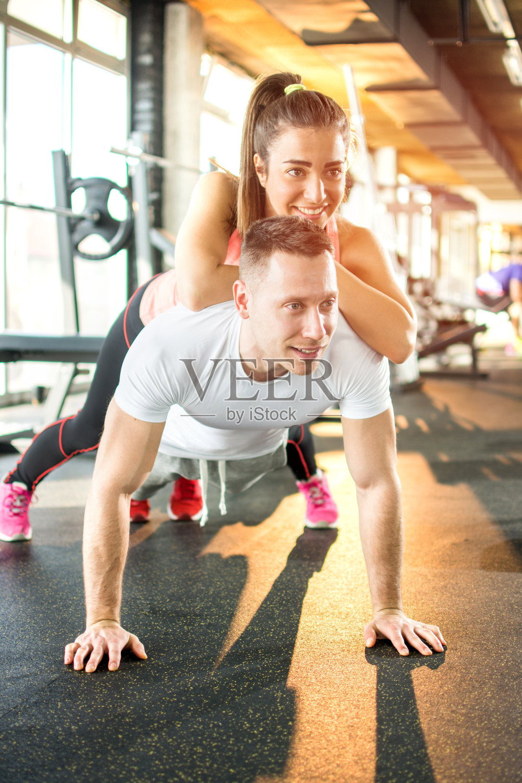 一对年轻的运动情侣在健身房一起锻炼。男人做俯卧撑，女孩躺在他身上。照片摄影图片