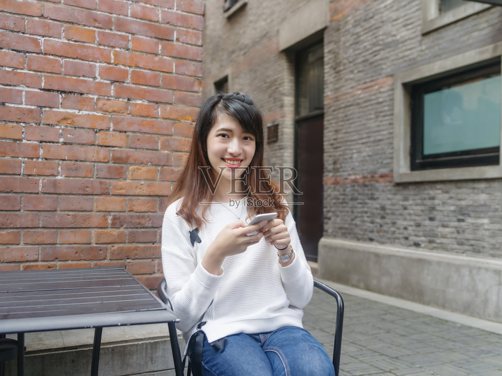 上海新天地，一名年轻美丽的中国女子穿着白色毛衣和蓝色牛仔裤坐在桌子前使用手机。照片摄影图片