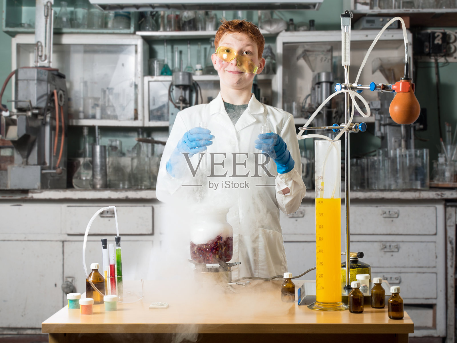 一个穿着白袍、戴着眼镜的男生站在化学桌子旁边，手里拿着烧瓶，背景是化学实验室，有桌子和化学设备照片摄影图片