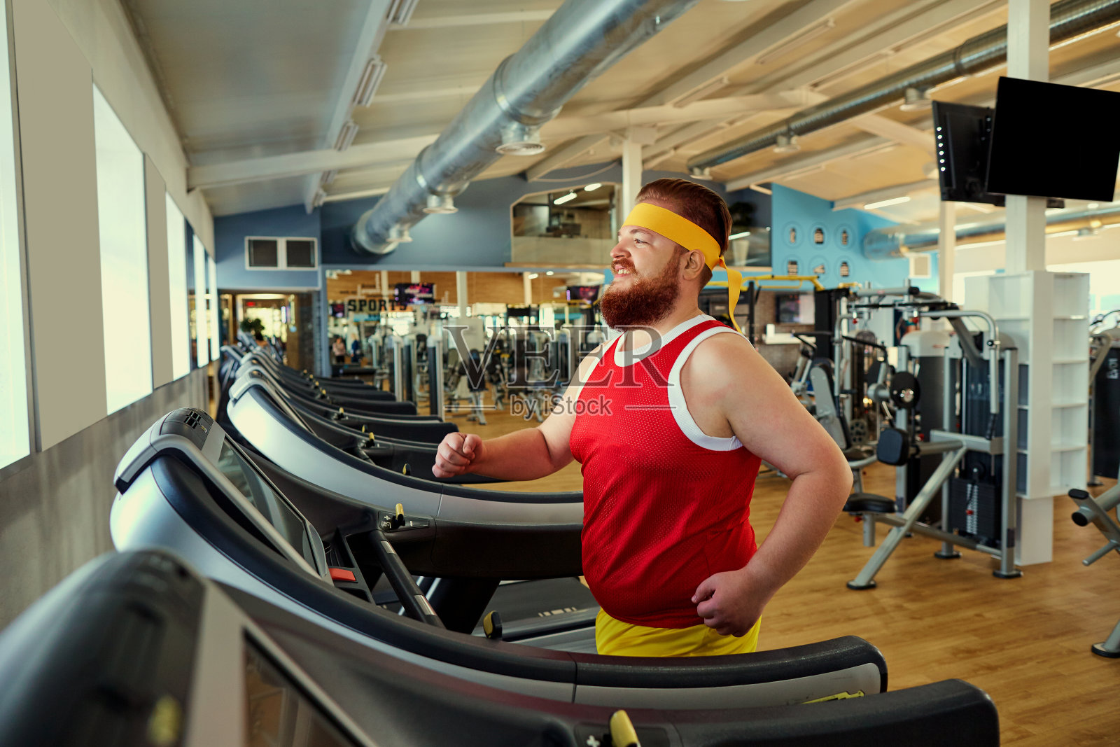一个胖子正沿着健身房的跑步机走。照片摄影图片
