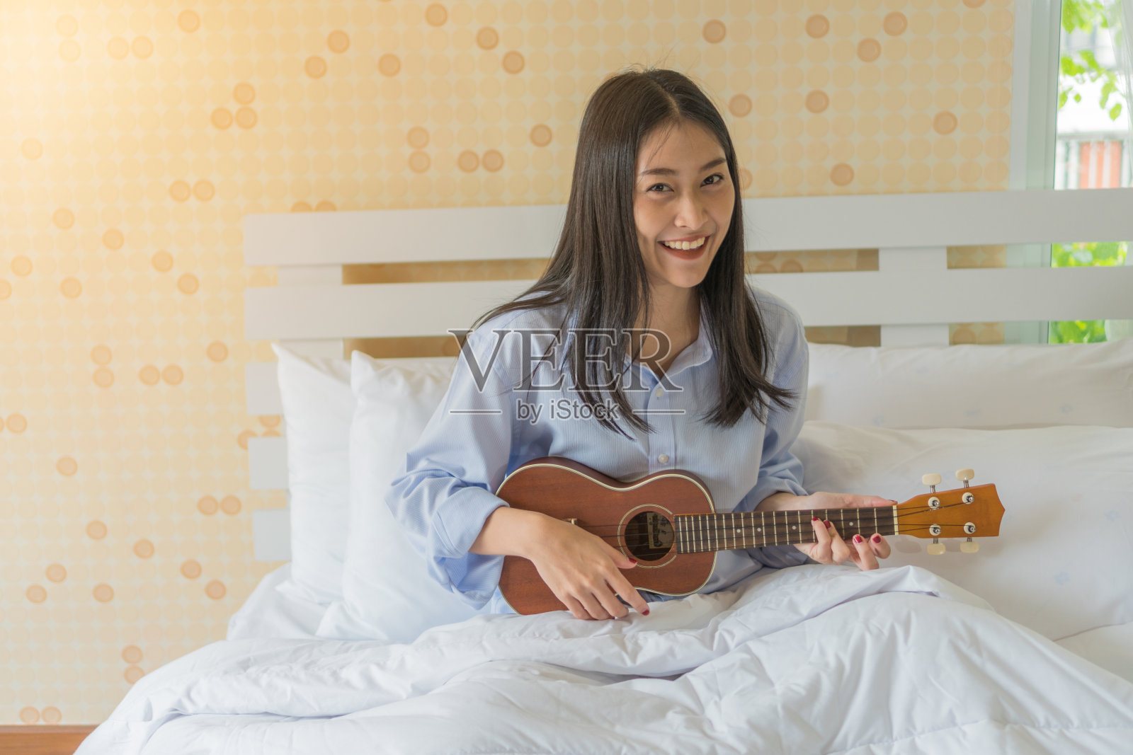 快乐微笑的亚洲女孩在床上弹奏尤克里里。照片摄影图片