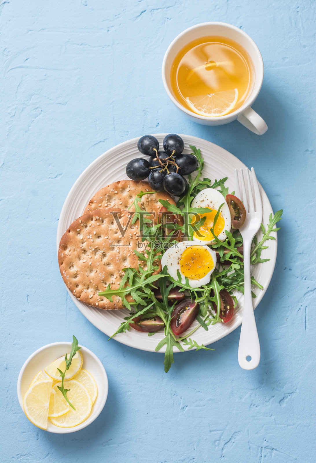 早餐碟-饼干，芝麻菜，圣女果，煮鸡蛋沙拉和绿茶与柠檬在蓝色背景，俯视图照片摄影图片