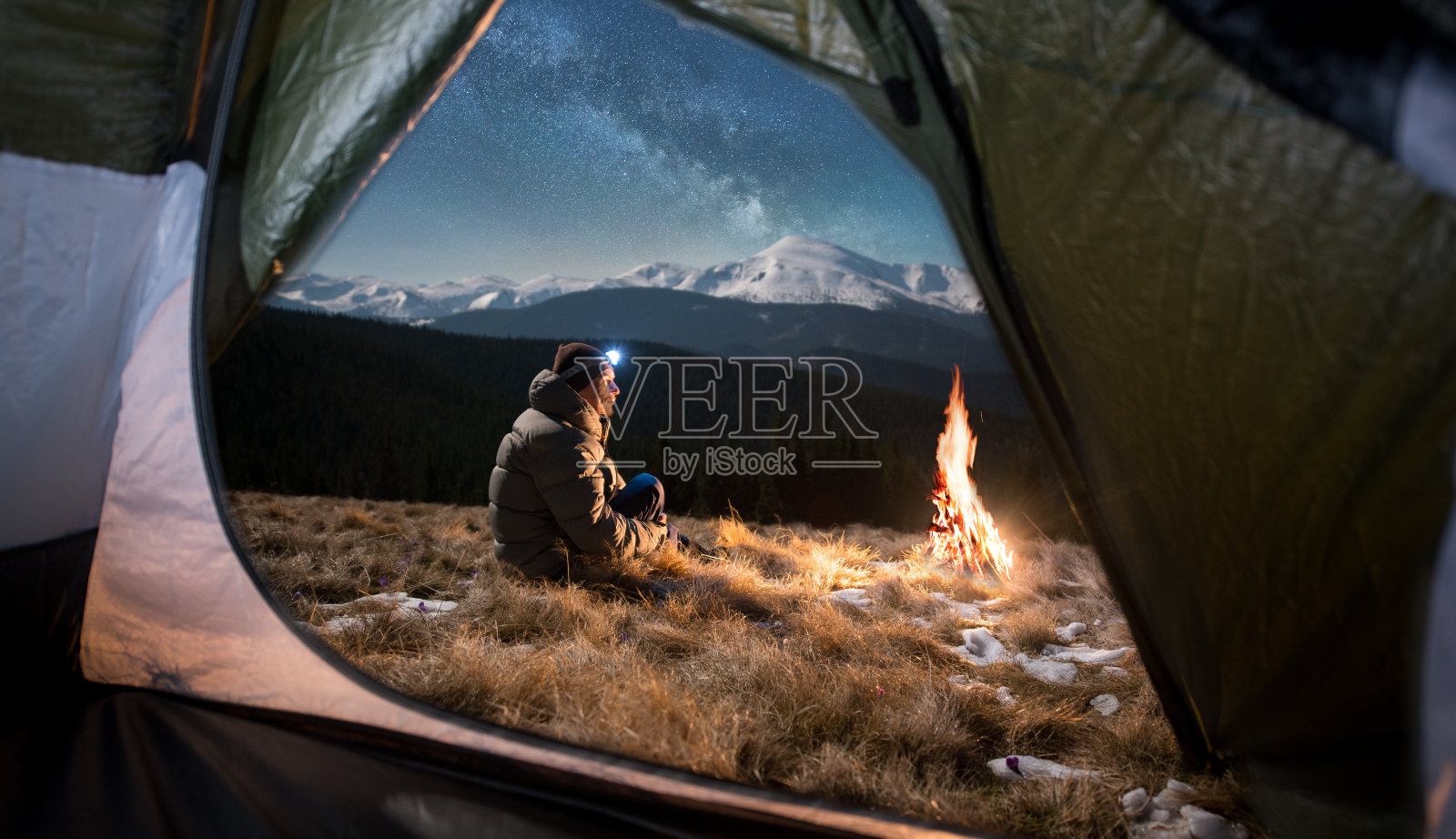 从帐篷里面望去，这位男游客正在他的帐篷里休息。在满是星星和银河的美丽夜空下，一个带着头灯的男人坐在篝火旁照片摄影图片