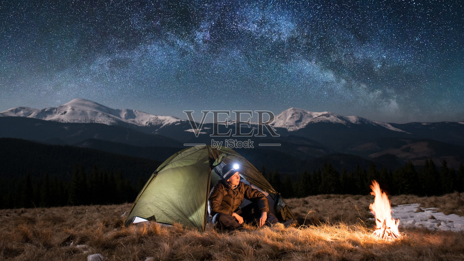 男游客晚上在山中露营休息。在满是星星和银河的美丽夜空下，一个带着头灯的男人坐在篝火和帐篷旁。图片宽高比16:9照片摄影图片