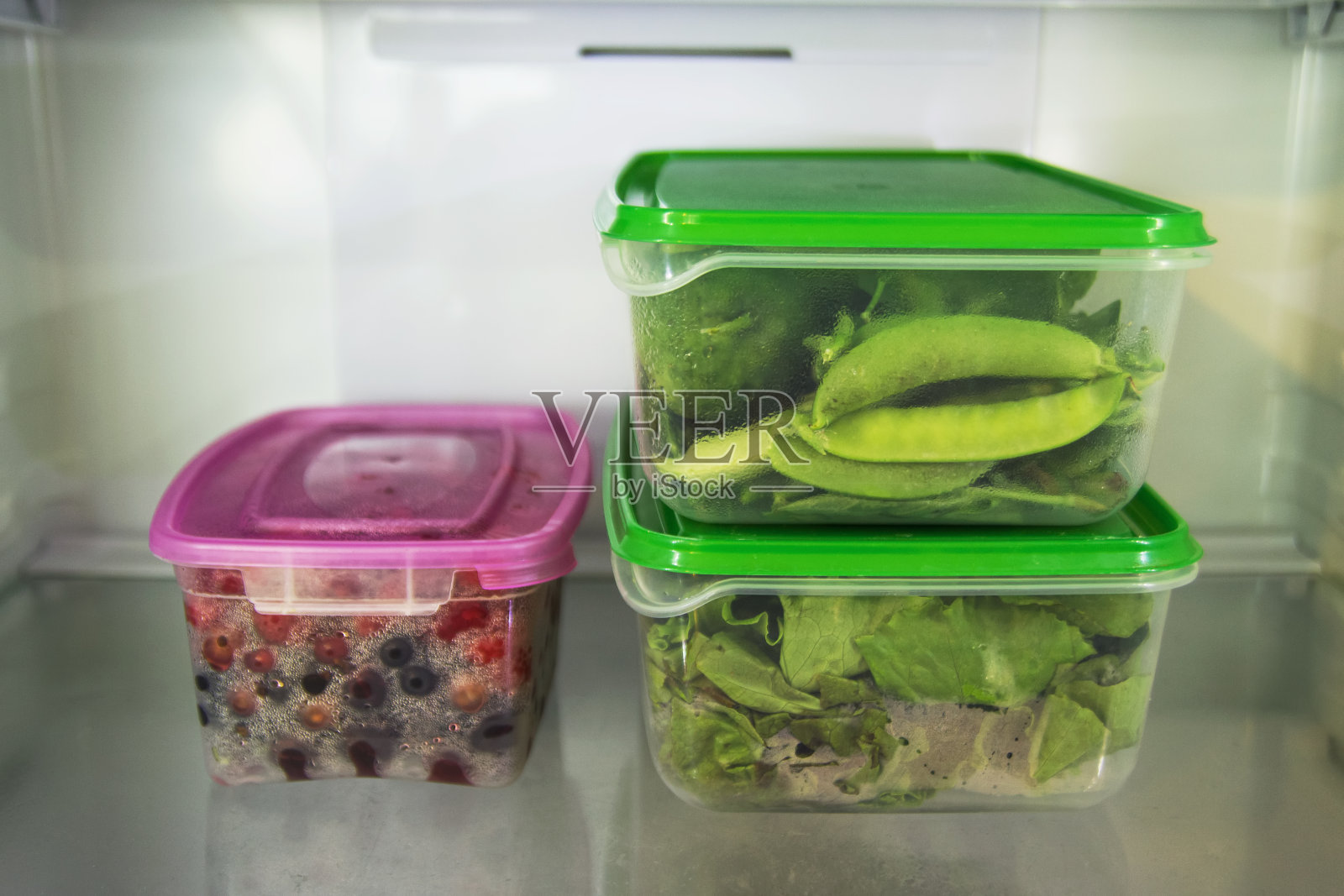 冰箱的架子上放着两个塑料食品容器，里面装着绿色蔬菜(沙拉、罗勒、莳萝、酢浆草、豌豆、黄瓜、胡椒)，另一个装着浆果(醋栗、覆盆子、醋栗)。照片摄影图片