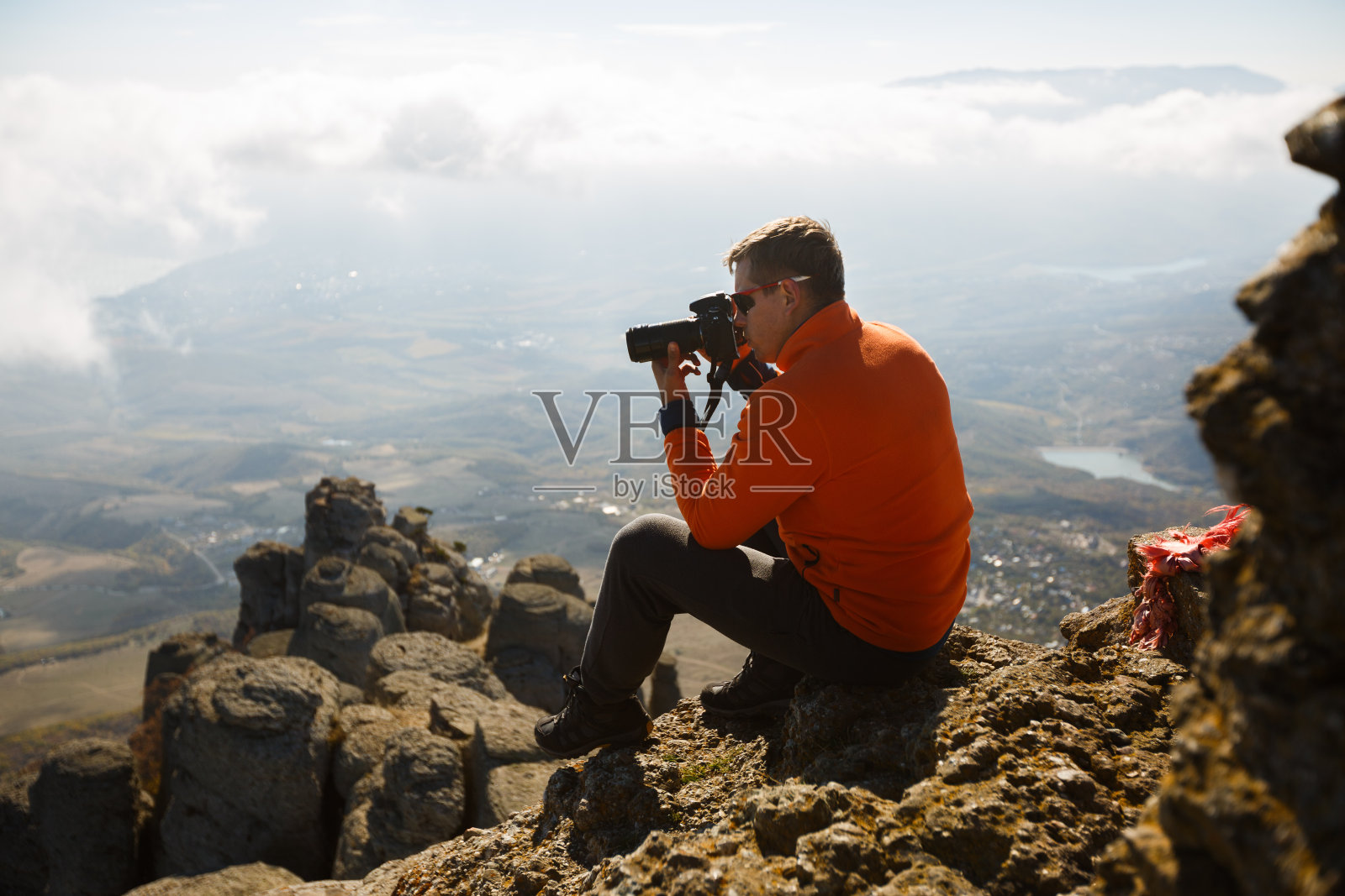 年轻的专业旅行家拿着数码单反相机拍摄户外美妙的山景。一个徒步旅行者坐在山顶的一块岩石上，在云层上摄影照片摄影图片
