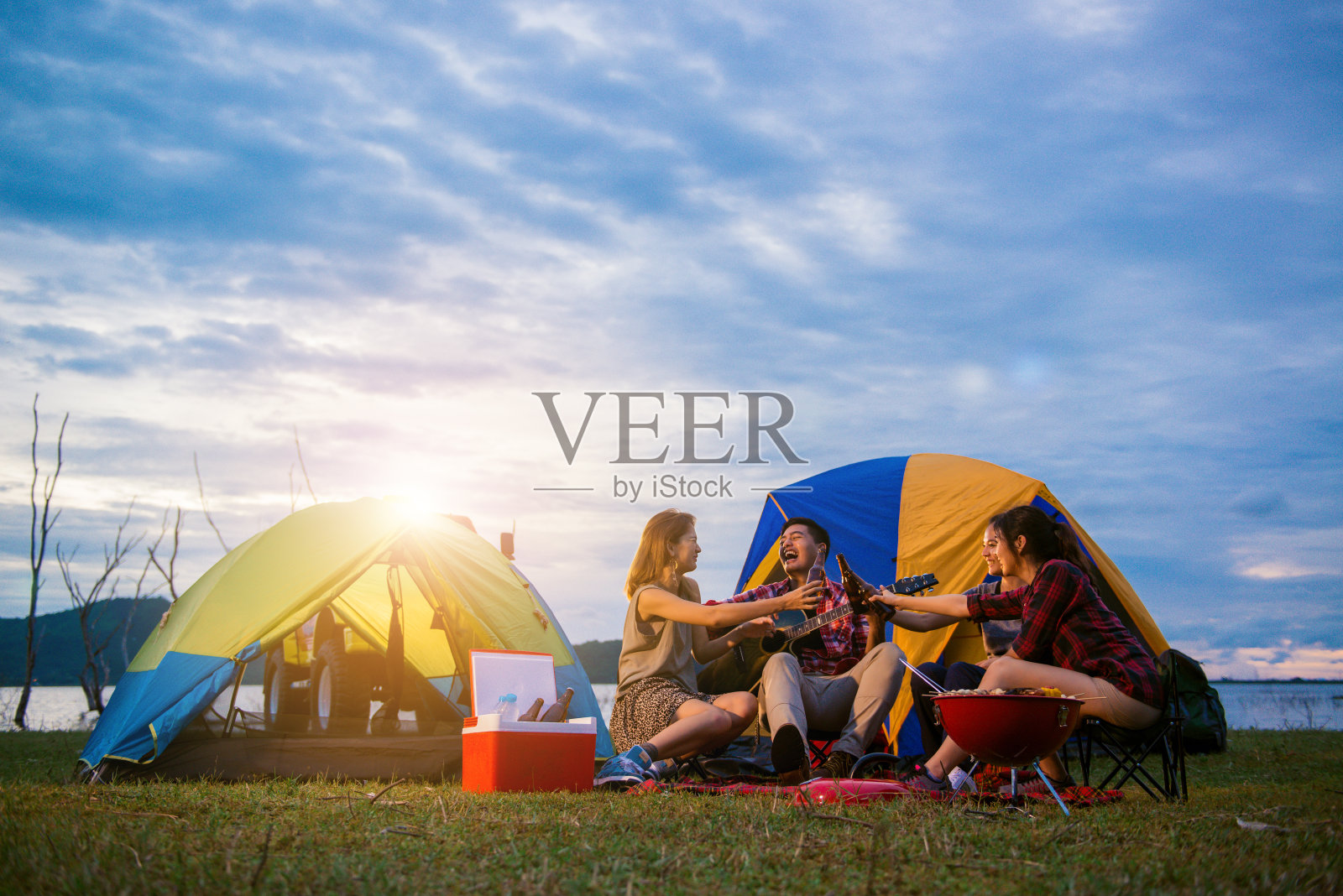 一群男人和女人喜欢在湖边露营野餐和烧烤，背景是帐篷。年轻的混血亚裔男女。年轻人的手为啤酒瓶干杯欢呼。照片摄影图片