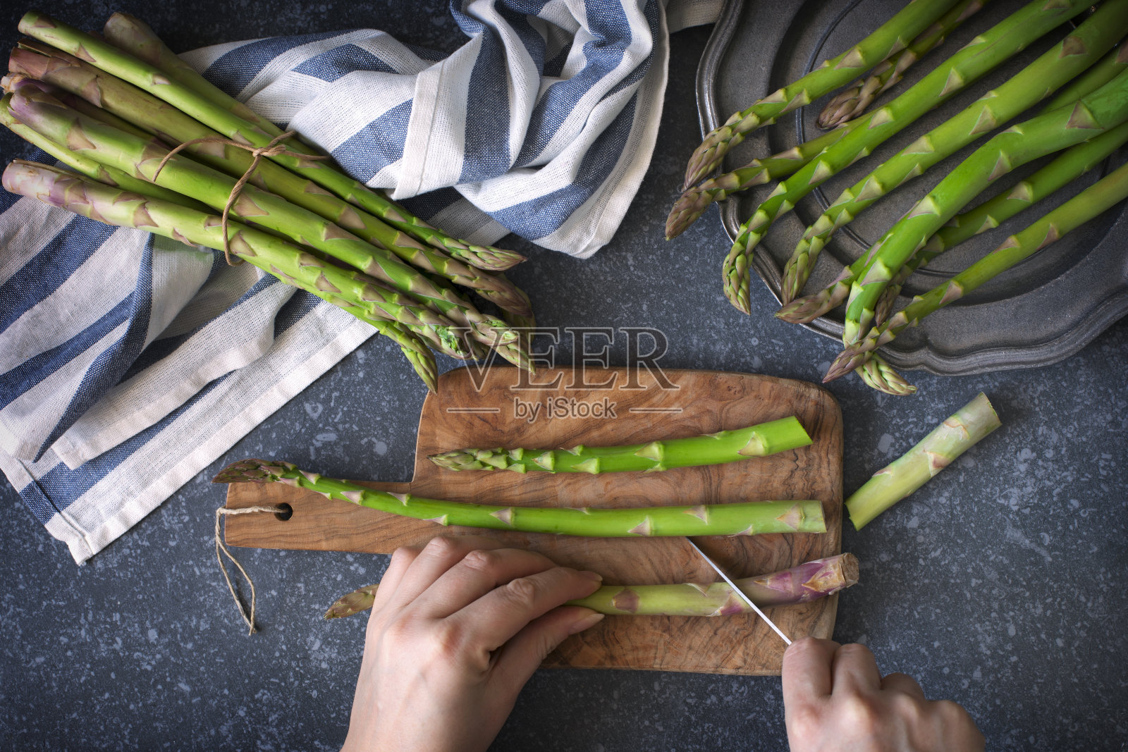 石头背景上的新鲜生芦笋。女人用女人的手在木制烹饪板上切芦笋。俯视图照片摄影图片