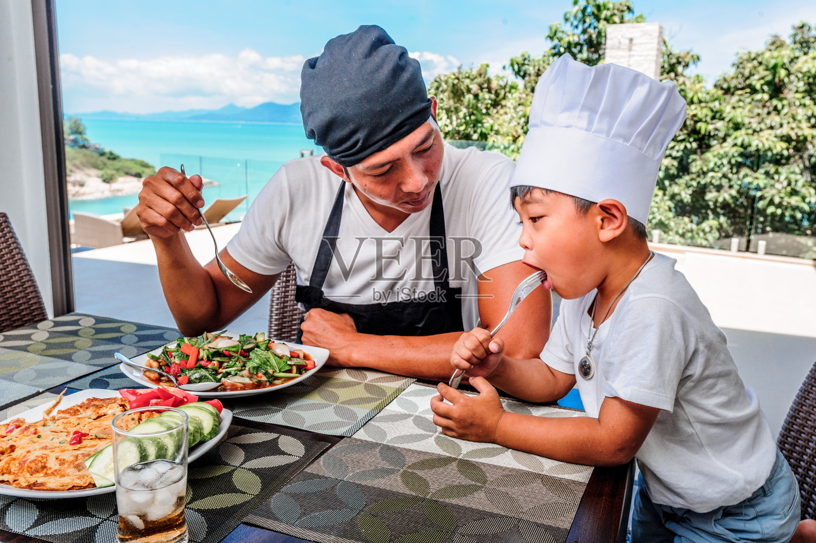 泰国男子和他可爱的儿子在家吃午饭。玩厨师游戏:围裙，厨师帽和泰国菜。令人惊叹的海景豪华别墅。鲜艳的颜色概念照片摄影图片