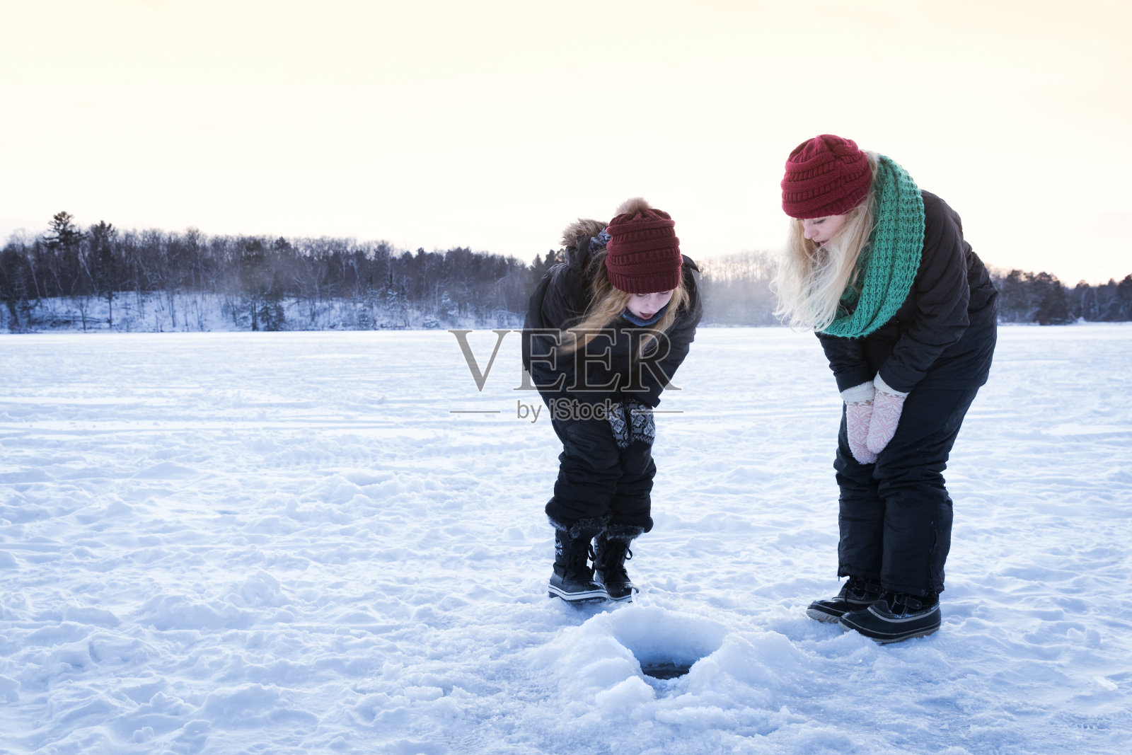 两个金发女孩看着结冰的湖面上的一个冰上钓鱼洞照片摄影图片