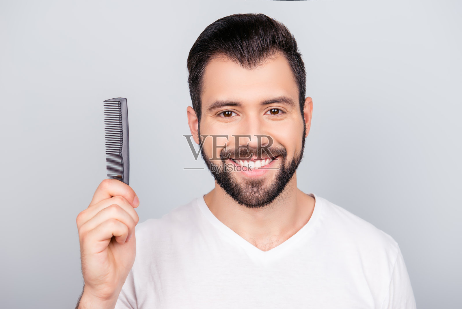 自信微笑英俊的年轻黑发胡子男子在白色t恤站在纯净的背景，拿着一把梳子。如此性感迷人，令人愉悦，时尚照片摄影图片