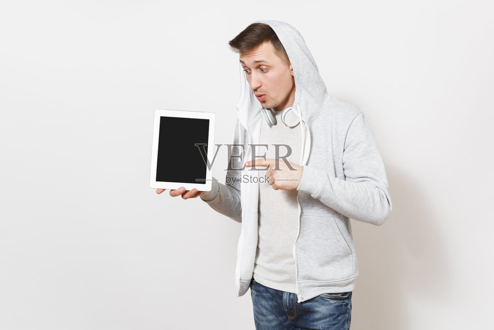 年轻英俊惊讶的学生在t恤和轻型运动衫兜帽与耳机手持平板电脑和指向它在工作室的白色背景。技术的概念照片摄影图片