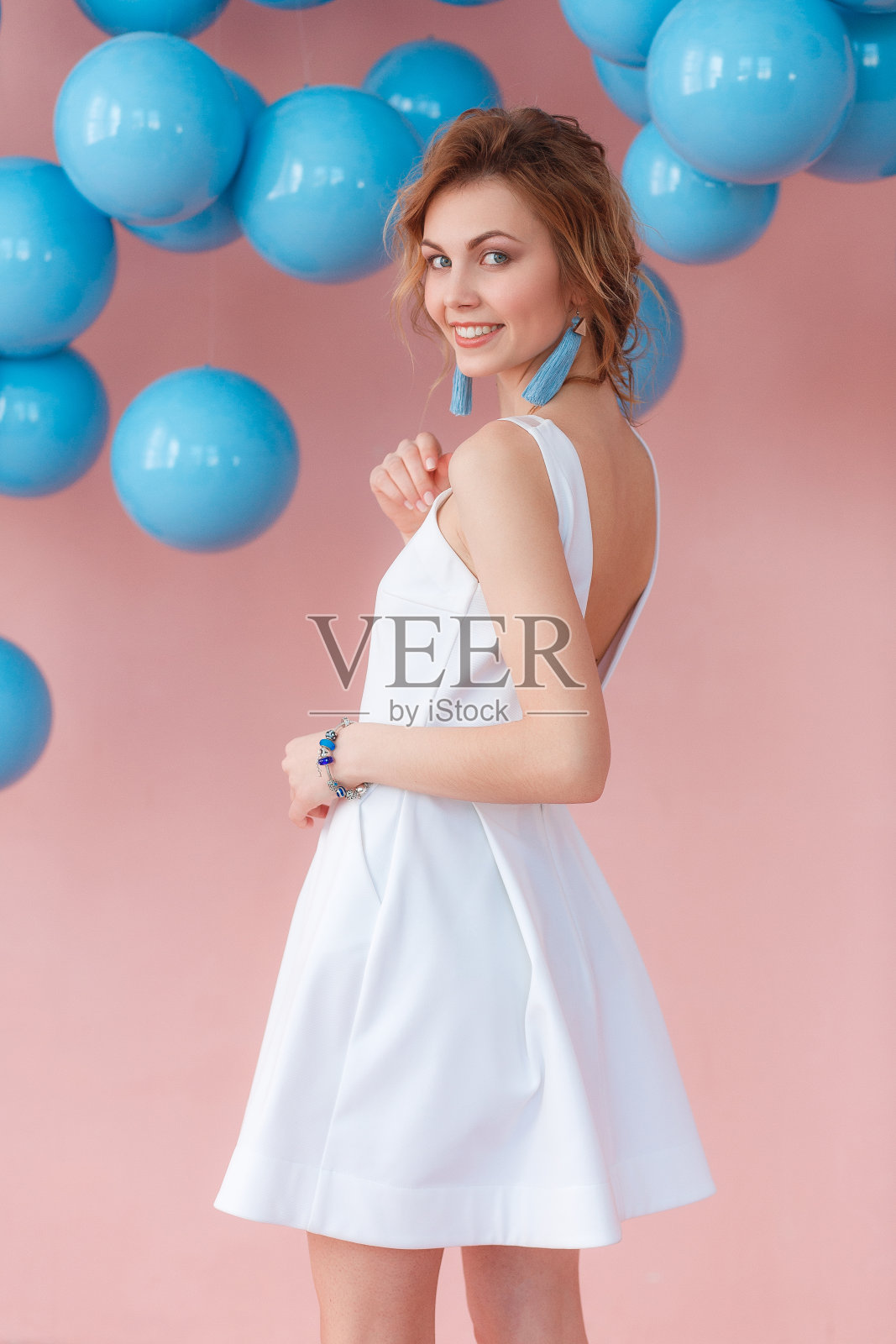 年轻女子在白色的鸡尾酒裙摆在粉红色的墙壁背景与蓝色的球悬挂照片摄影图片