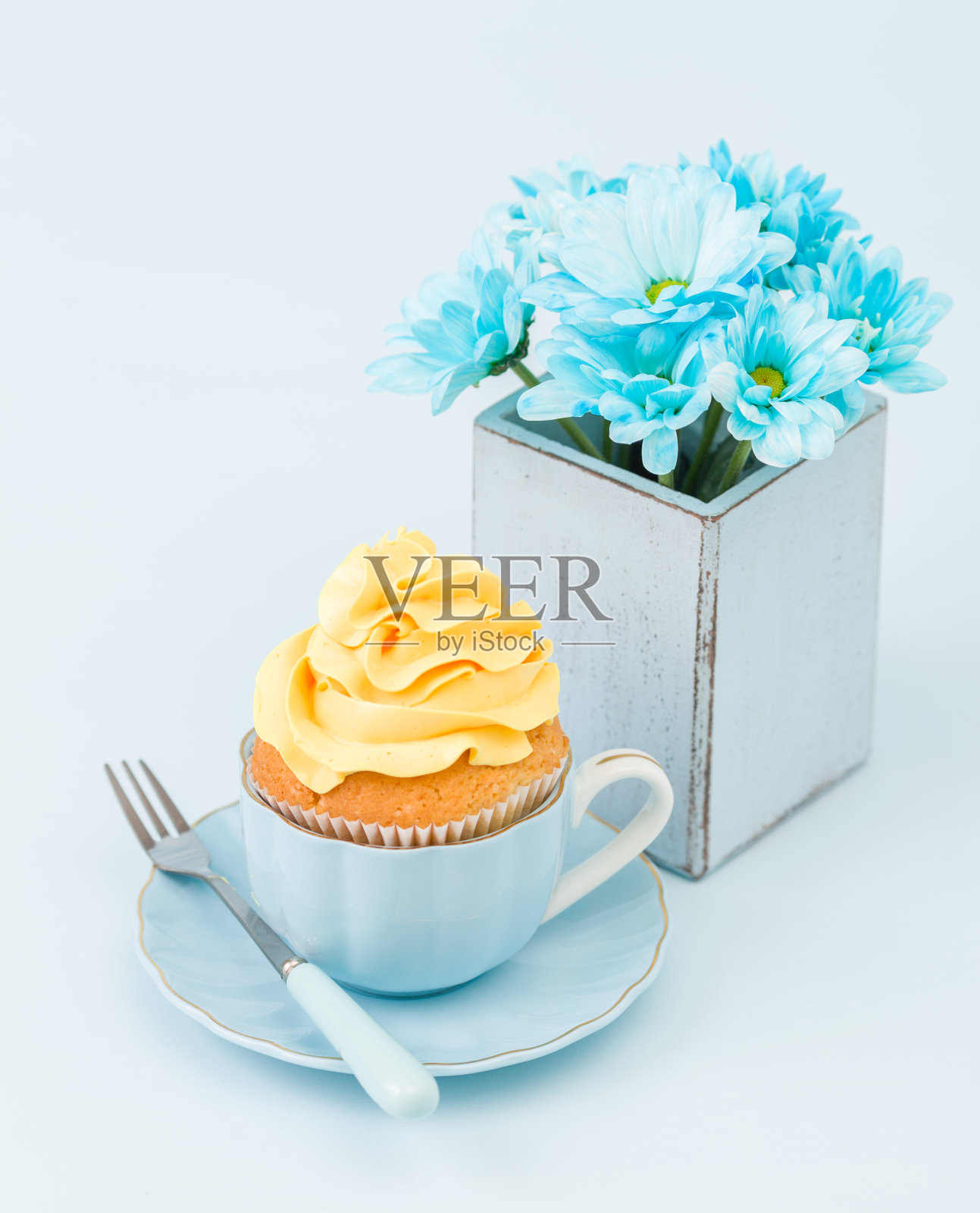 纸杯蛋糕与嫩黄奶油装饰和蓝色菊花花束复古破旧别致的花瓶在蓝色的粉彩背景。照片摄影图片