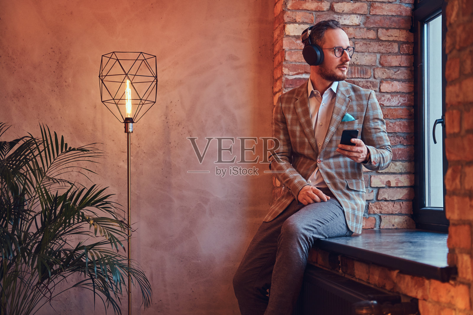 一个穿着法兰绒西装、戴着眼镜的时尚男士的肖像，他坐在一个有阁楼的房间的窗台上，用手机和耳机听音乐。照片摄影图片