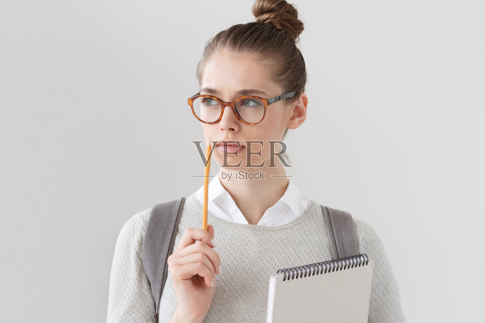 特写的好看的年轻女人孤立地站在灰色的背景，戴眼镜，拿着铅笔旁边的嘴唇，思考和看左边，试图找到答案或解决方案。照片摄影图片
