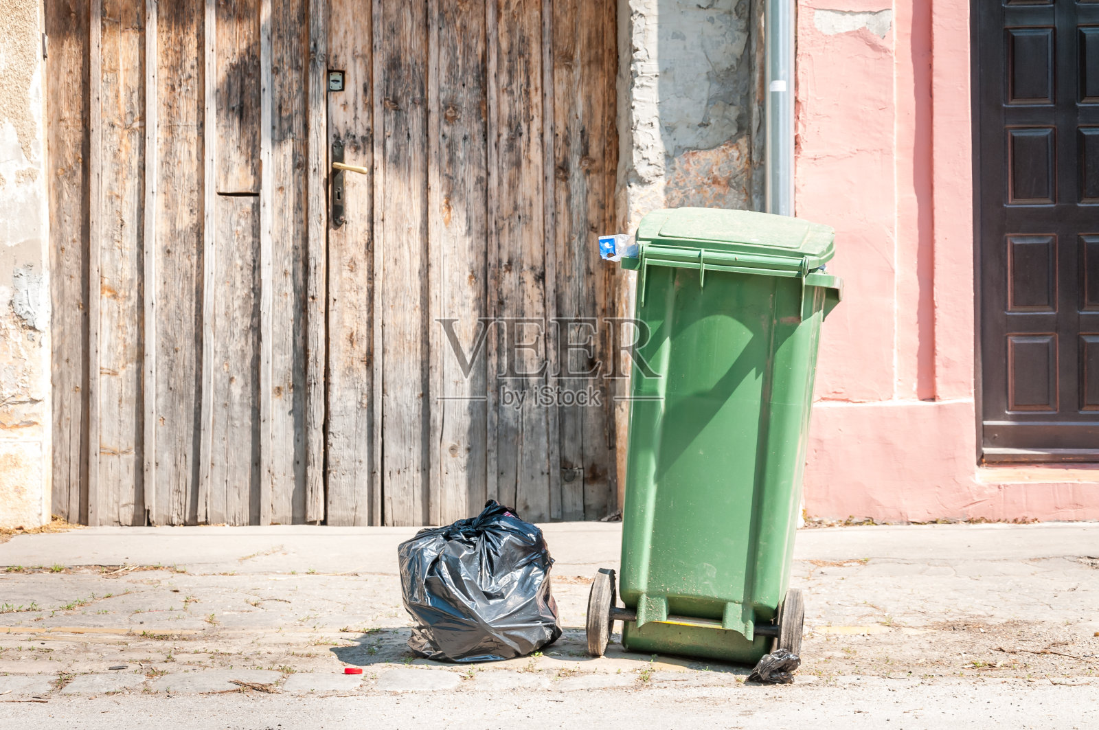 一个绿色的垃圾桶和黑色的塑料垃圾袋在城市的街道上，等待倾卸卡车收集在房子前面照片摄影图片