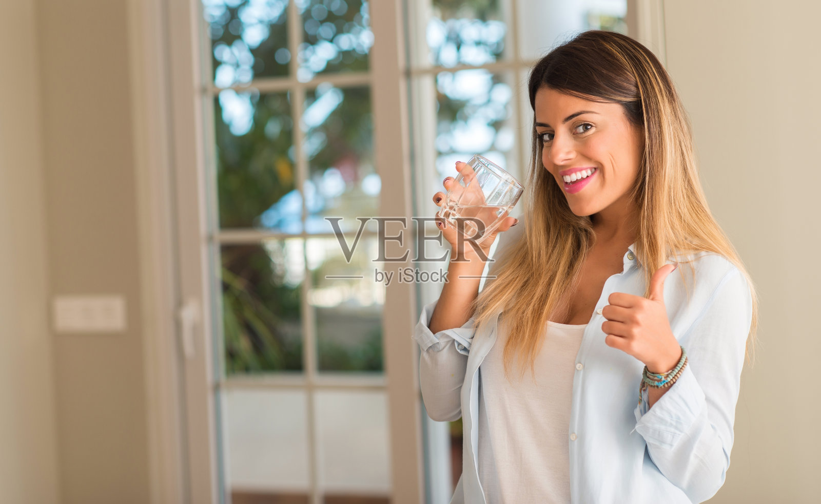 美丽的年轻女子喝一杯水，并在家里竖起大拇指。生活方式的概念。照片摄影图片
