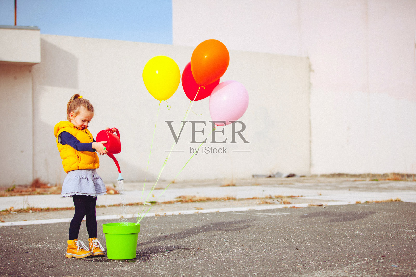 可爱的金发小女孩被混凝土墙包围着，正在给彩色气球播种和浇水照片摄影图片