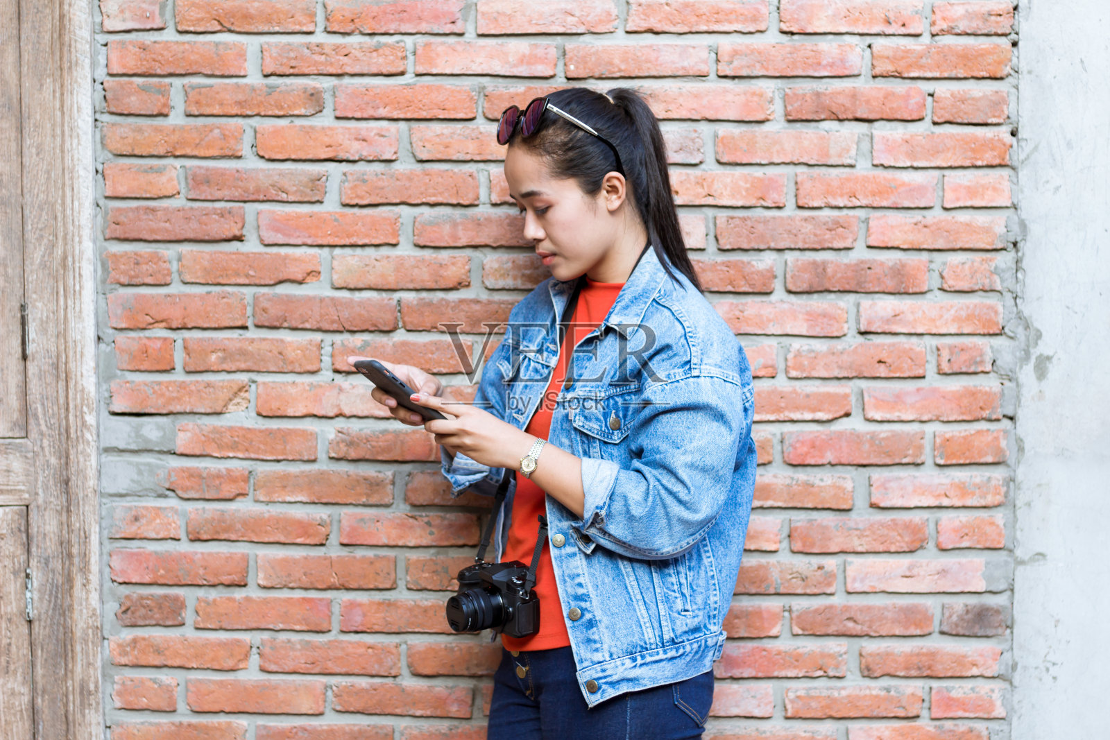 一个头戴墨镜的女人，穿着夹克、牛仔裤和橘色衬衫，靠墙站着玩手机和相机。照片摄影图片