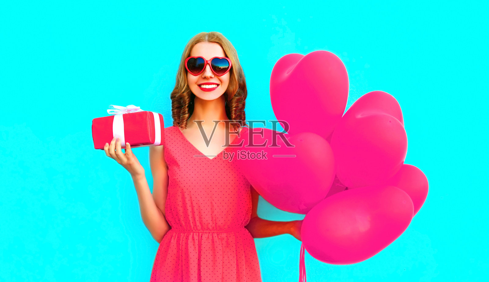 快乐微笑的年轻女子手持礼品盒粉红色的心形气球在彩色的蓝色背景照片摄影图片