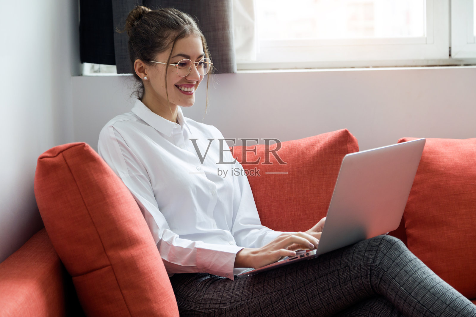 微笑的年轻女商人坐在酒店房间的沙发上用她的笔记本电脑工作。照片摄影图片
