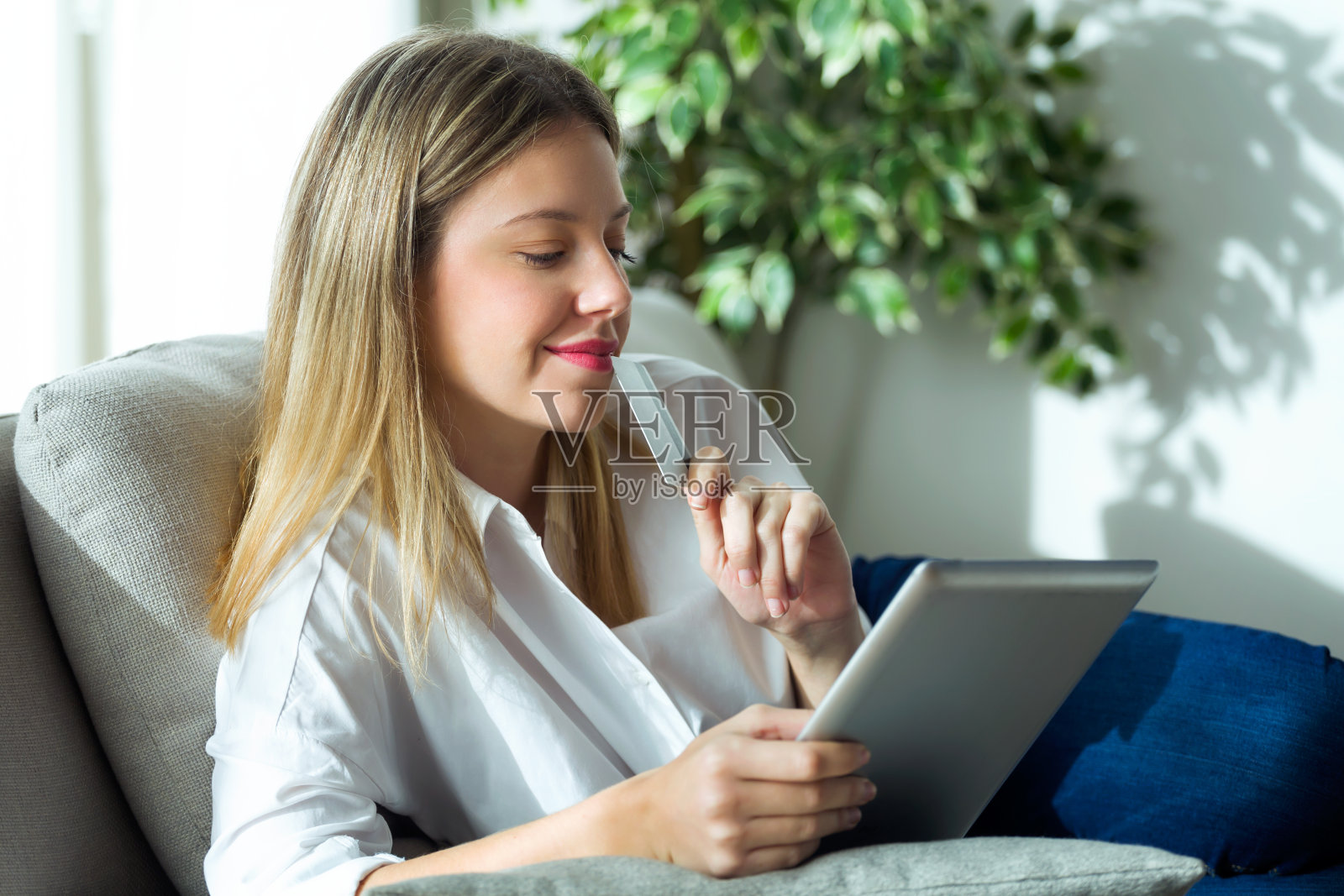 自信的年轻女子拿着白色的信用卡，坐在家里的沙发上用电脑在线购物。照片摄影图片