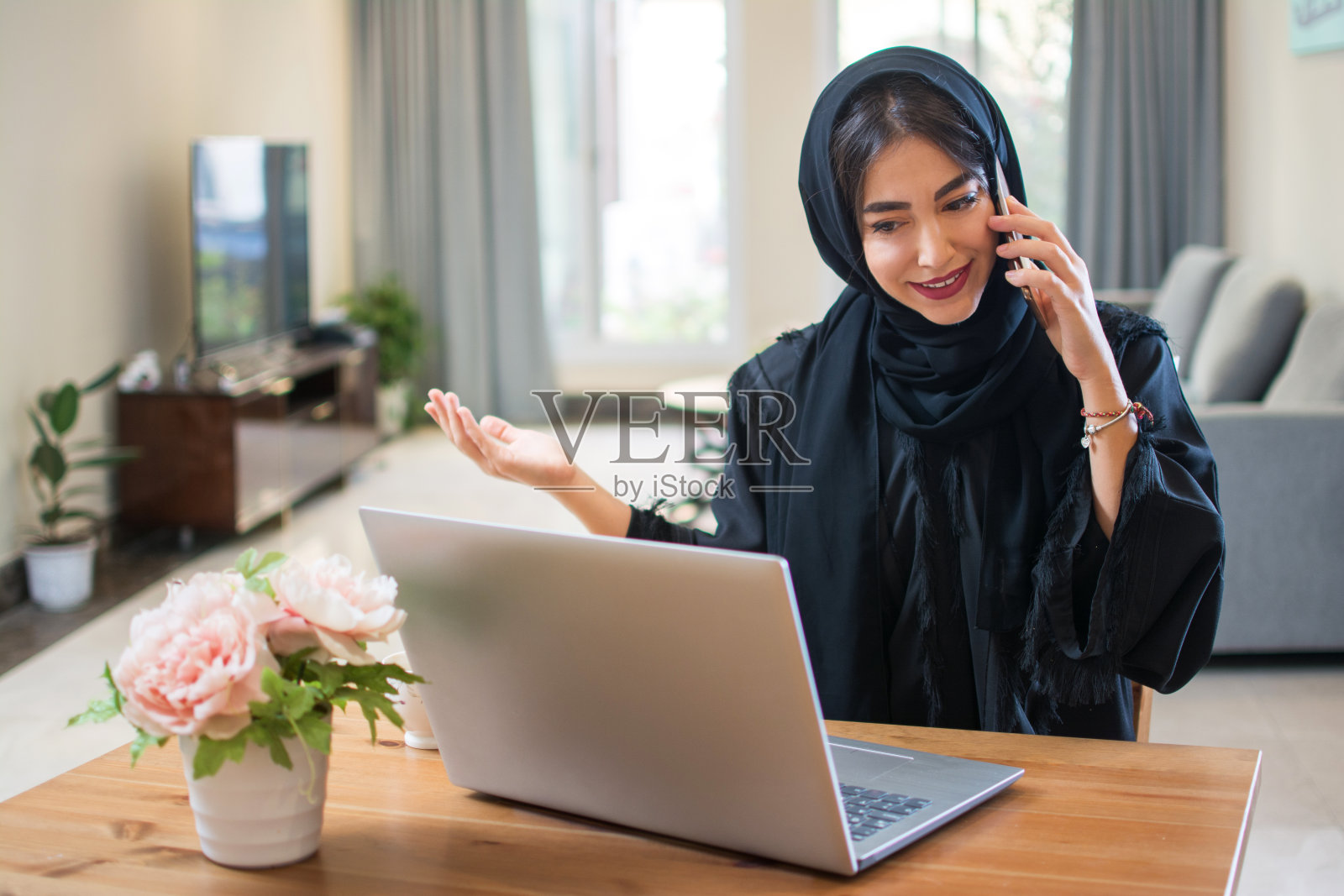 美丽的阿拉伯年轻女子在阿拉伯通过手机交谈，而坐在附近的笔记本电脑在她的豪华住宿照片摄影图片
