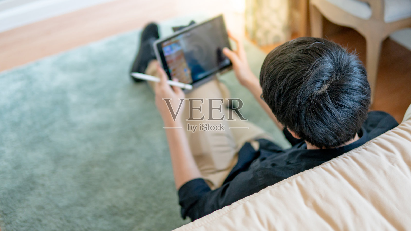 在舒适的卧室里，年轻的亚洲商人坐在床边的地毯上使用数码平板电脑和钢笔。具有现代电子小玩意概念的家居生活方式照片摄影图片