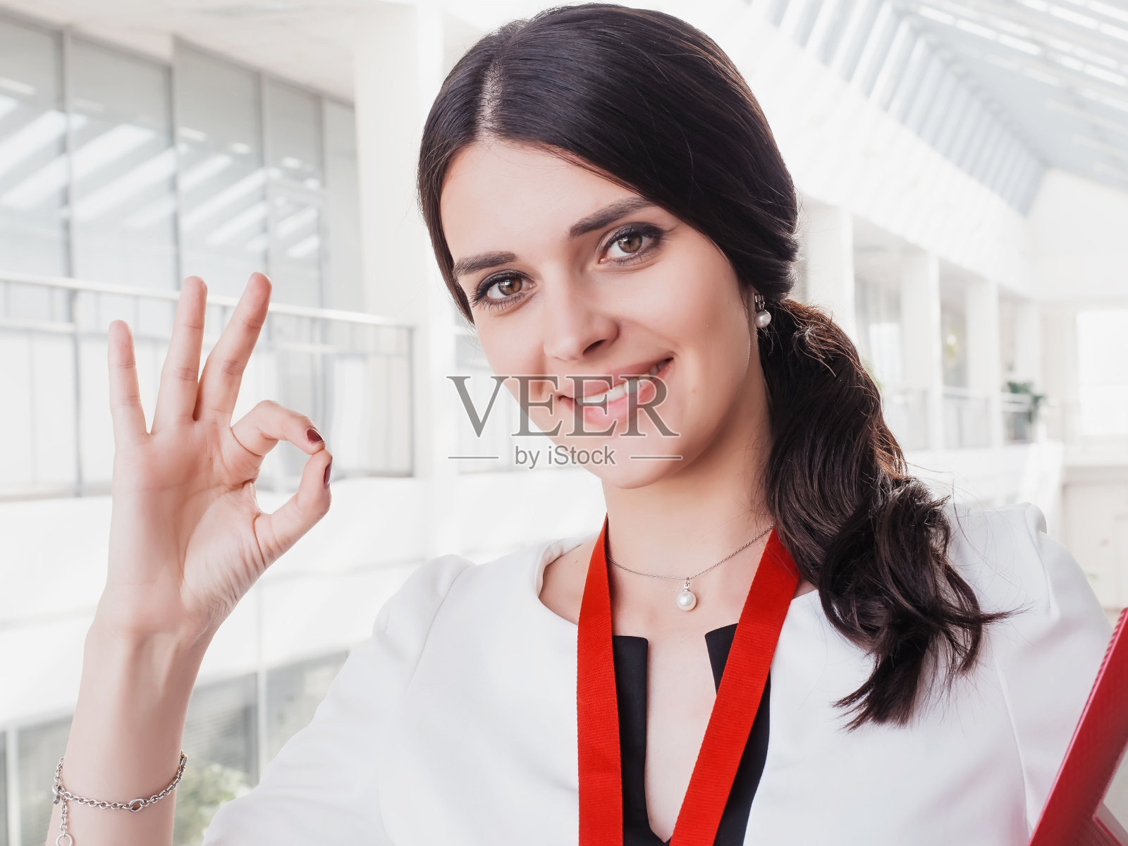 年轻微笑的女孩做了成功的工作显示手势大拇指向上。美丽微笑的女商人站在白色的办公室背景。手里拿着文件夹的女商人照片摄影图片