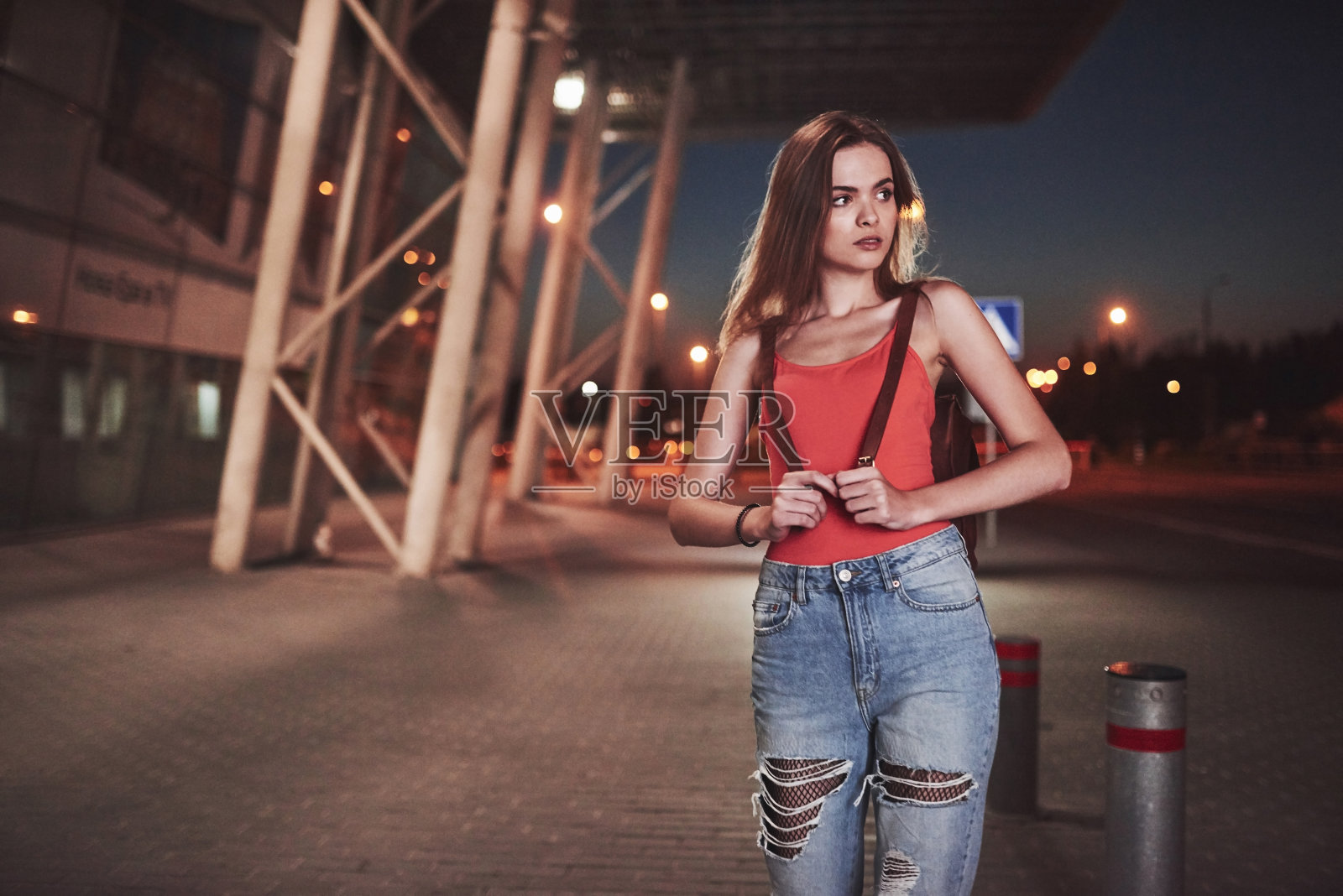 在一个温暖的夏夜，一个年轻美丽的女孩背着一个背包站在机场或火车站附近的街道上。她刚到，等出租车或她的朋友照片摄影图片