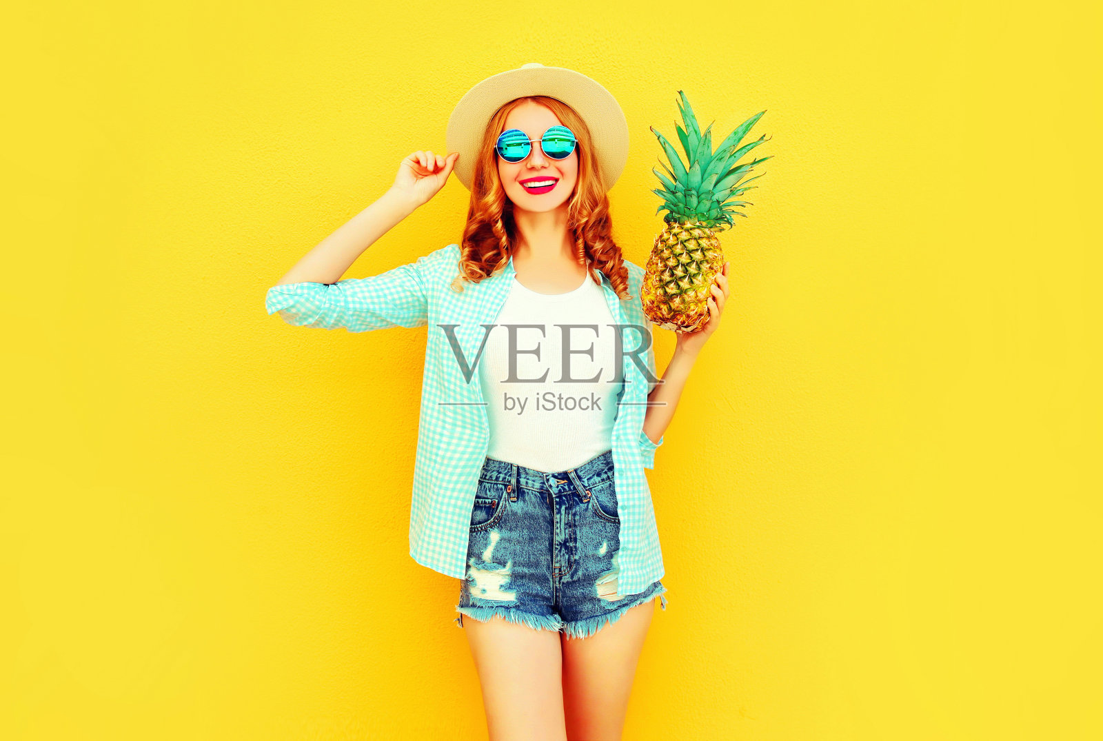 肖像快乐的微笑女人与菠萝有乐趣在夏天草帽，太阳镜，短裤上彩色的黄色背景照片摄影图片