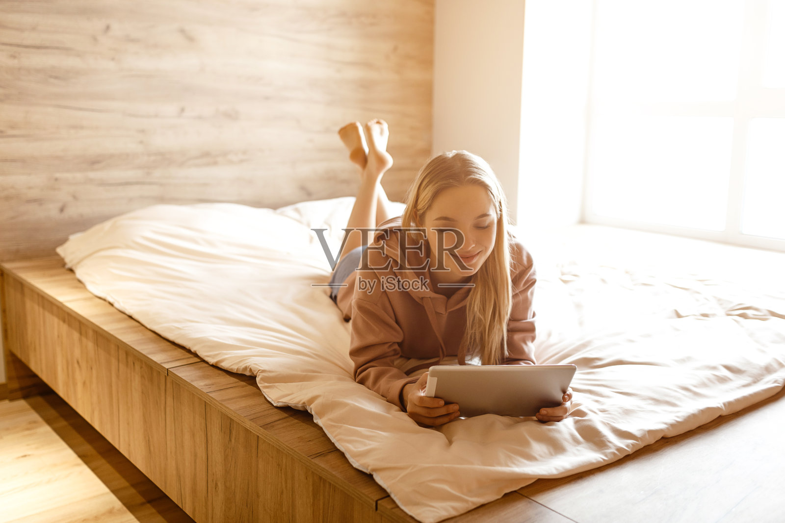 年轻漂亮的金发女人今天早上躺在床上。她手里拿着笔记本电脑，看着它。冷静平和、集中的模式。阳光从窗外照进来。照片摄影图片