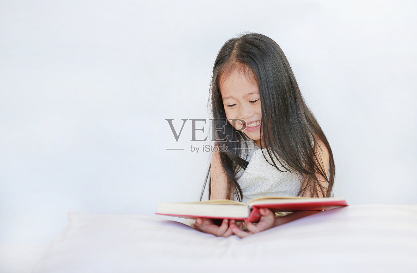 微笑的小亚洲女孩读精装书与枕头躺在床上对白色的背景。照片摄影图片