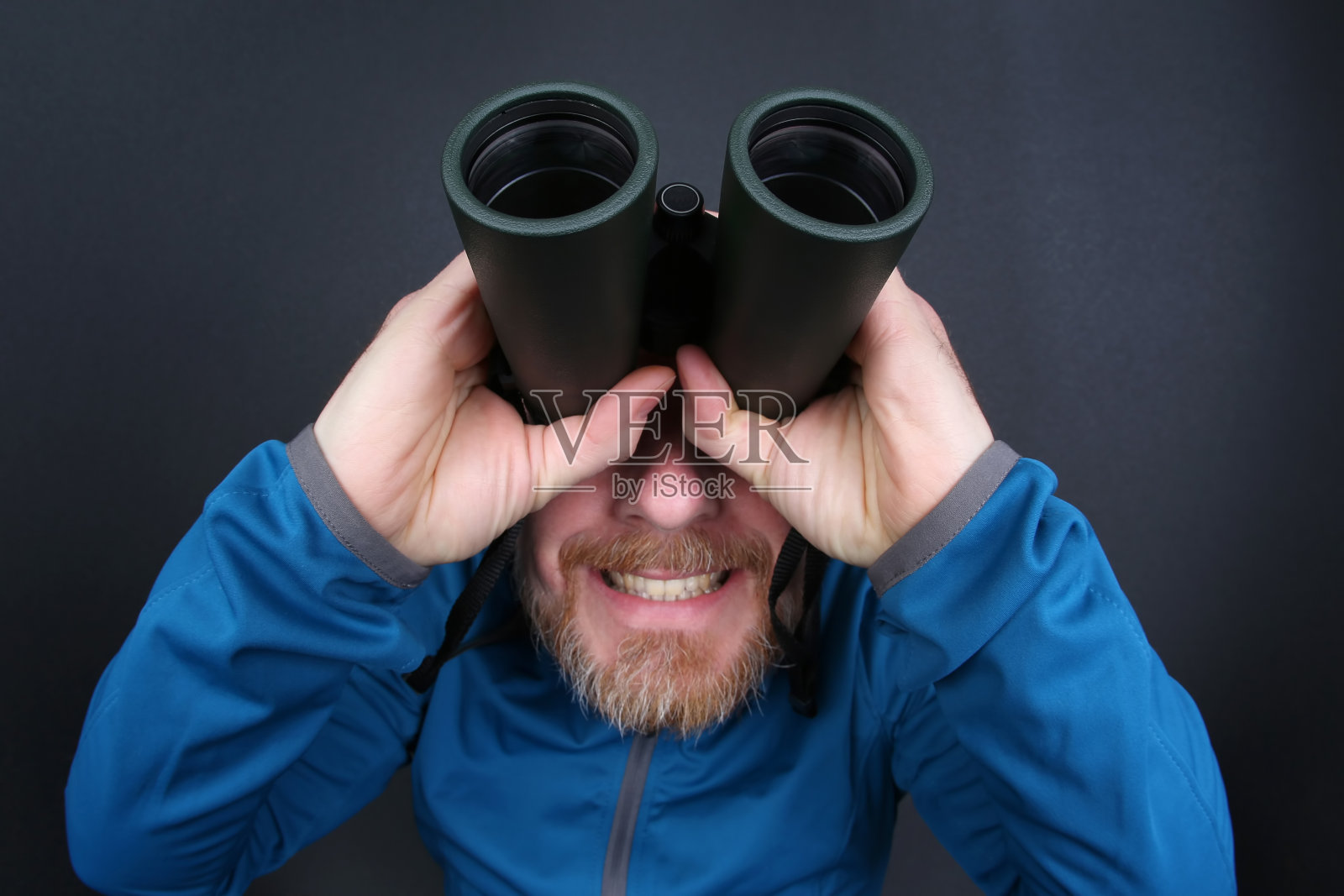 在灰色的背景下，蓄着胡子的人用双筒望远镜抬头看照片摄影图片