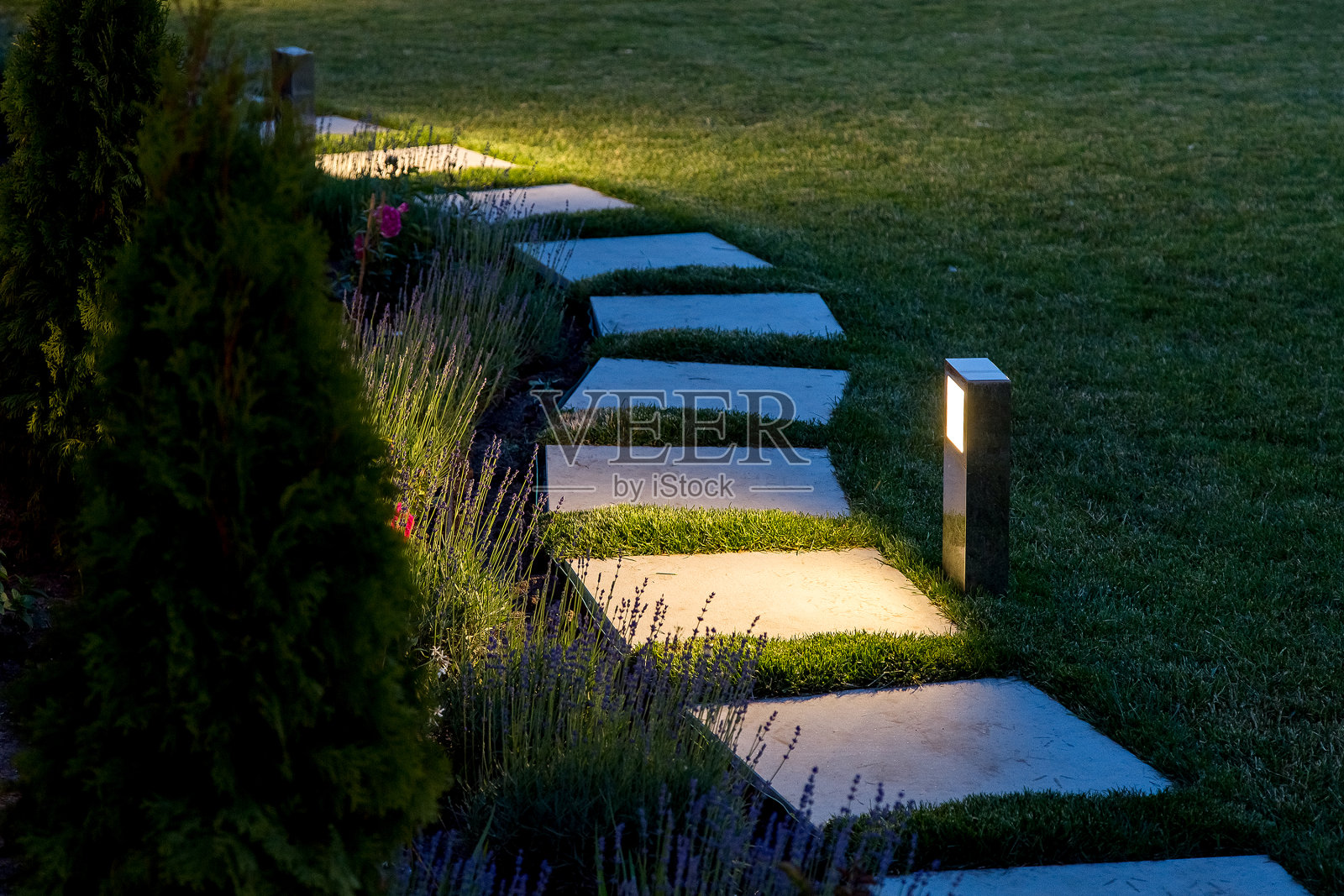 在带有花坛和草坪拷贝空间的后院花园中，一盏温暖的灯笼照亮了方形瓷砖的大理石路径。照片摄影图片