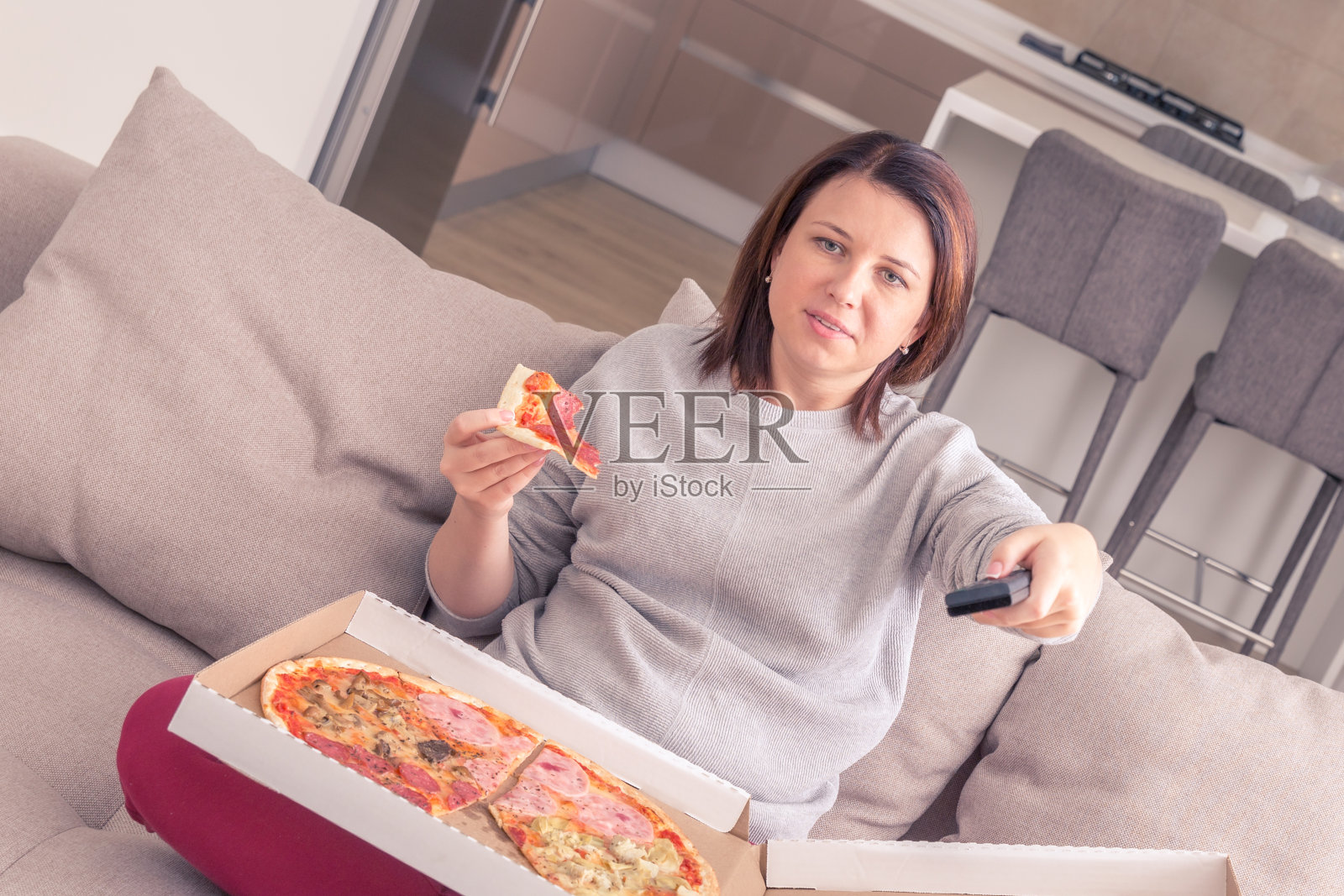 漂亮女人在家里一边吃披萨一边看电视一边拿着遥控器，橙色调照片摄影图片