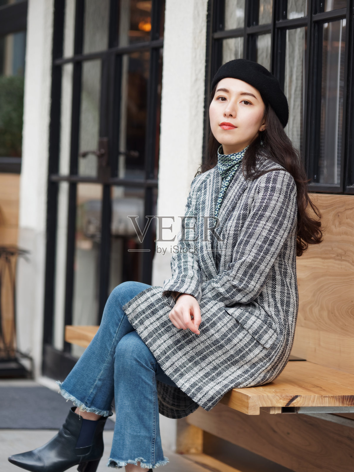 美丽的中国年轻女子穿着外套和牛仔裤坐在户外咖啡馆，看着镜头与腿交叉，自信的商业女性，户外时尚肖像魅力时尚女士。照片摄影图片