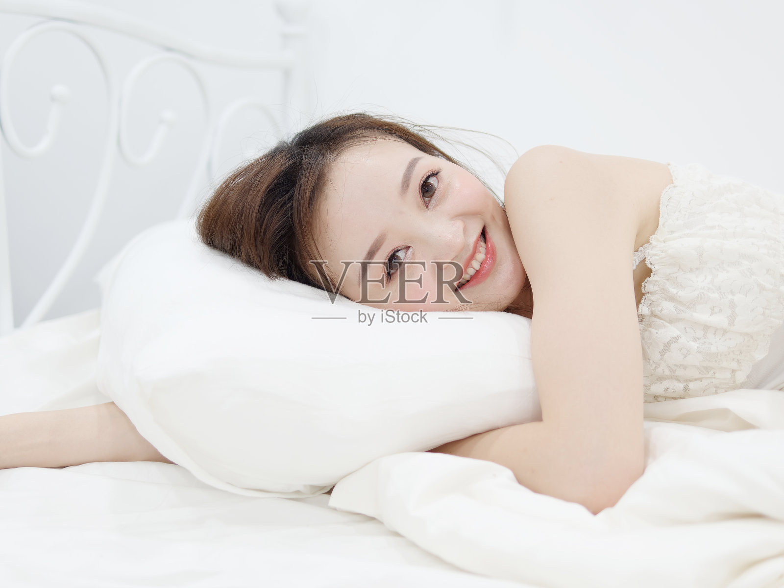美丽的女人穿着白色的睡衣躺在床上，手臂放在枕头下对着镜头微笑。照片摄影图片
