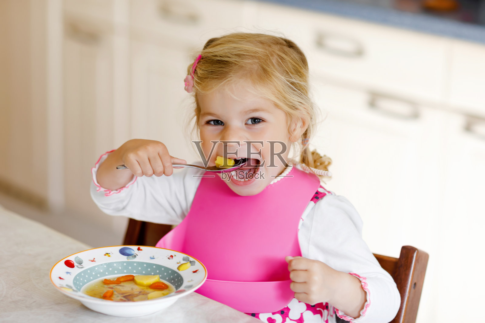 可爱的蹒跚学步的小女孩午餐吃健康的蔬菜，土豆和胡萝卜汤。可爱快乐的宝宝在家里或托儿所或幼儿园吃东西和学习用勺子。照片摄影图片