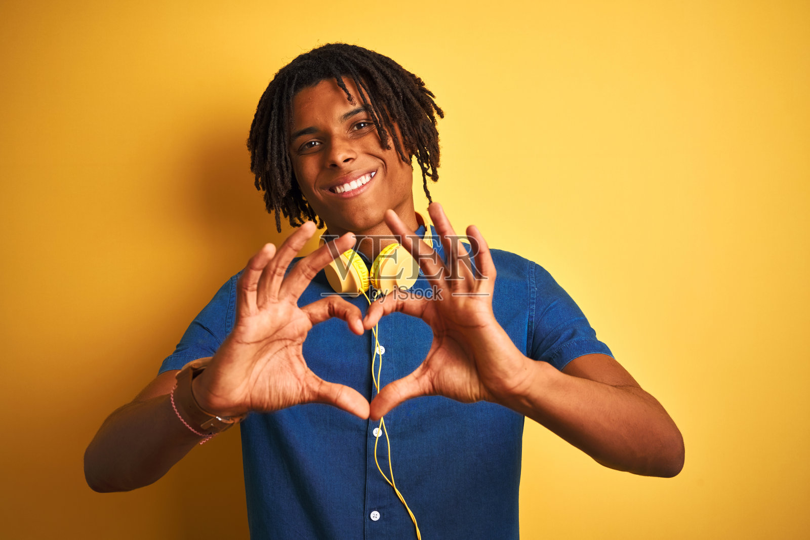 非洲裔美国人与发辫戴耳机在孤立的黄色背景微笑在爱显示心形符号和形状的手。浪漫的概念。照片摄影图片